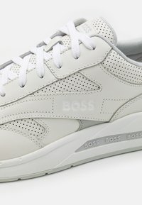 Bílé kožené sneakers s perforovanými akcenty a šedými detaily. Obsahují ploché bílé tkaničky a embosované logo na boku a podrážce.