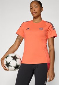 Oranje sportshirt met blauwe accenten en logo, gecombineerd met zwarte legging. Model houdt een witte voetbal vast met zwart sterpatroon.