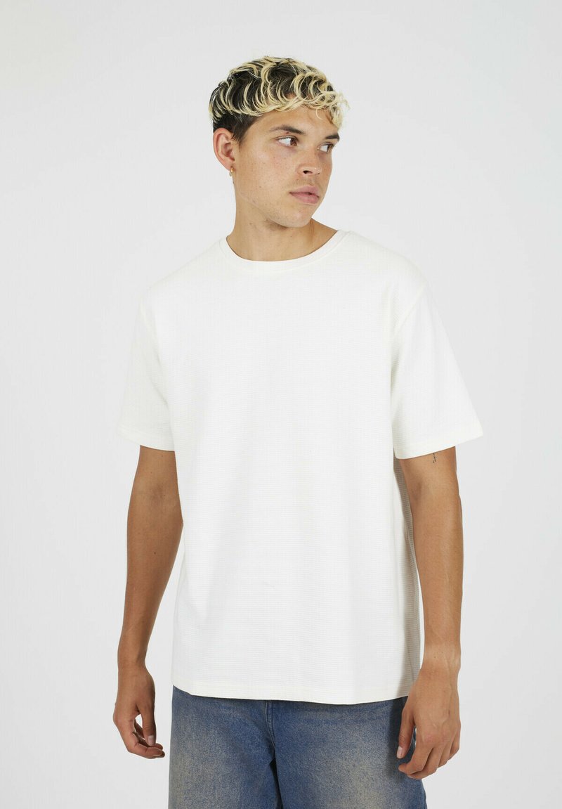 Młody mężczyzna o krótkich, kręconych blond włosach, ubrany w jednolity biały t-shirt z krótkim rękawem i niebieskie dżinsy, patrzący w prawo.