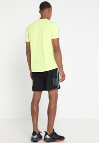 Camiseta de manga corta amarilla neón, pantalones cortos negros con una raya turquesa, zapatillas deportivas negras con detalles rojos, vistas desde atrás.