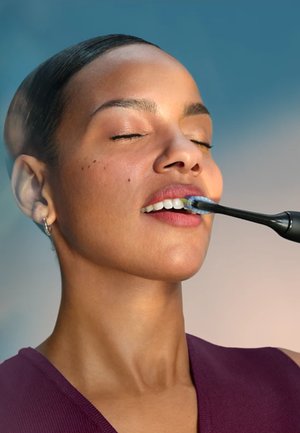 Eine Frau mit geschlossenen Augen putzt ihre oberen Zähne mit einer schwarzen elektrischen Zahnbürste vor einem unscharfen Hintergrund.