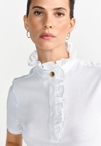 Beli zgornji del z volančastim ovratnikom, okrasnim gumbom, kratkimi rokavi in gladko teksturo tkanine. Preprost in eleganten dizajn.