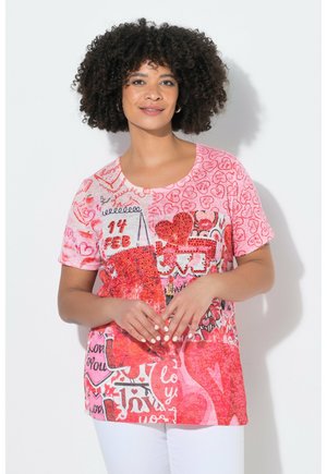 Kvinde med krøllet hår iført en lyserød og rød Valentinsdag-tema skjorte med hjerter, "14 FEB" og kærlighedsrelateret tekst, kombineret med hvide bukser.