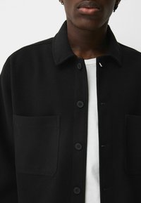 Czarna wełniana koszula-ramoneska z zapięciem na guziki z przodu, dwoma przednimi kieszeniami oraz klasycznym kołnierzem. Noszona z białym T-shirtem pod spodem.