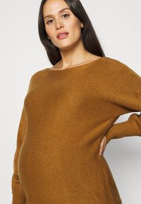 Strikket gravidsweater i varm brun, med bred halslinning og løs passform, samt teksturert ribbing på ermene og sidene.