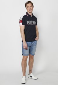 Sötétkék pólóing fehér és szürke díszítéssel, mellkasi logóval, roll-up farmer sorttal és fehér sportcipővel párosítva.