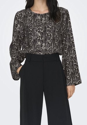 Woman wearing a long-sleeve, black and gray snake print blouse tucked into high-waisted, wide-leg black trousers with hands in pockets.