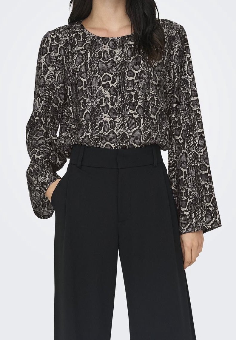 Femme portant un chemisier à manches longues imprimé serpent noir et gris, rentré dans un pantalon noir taille haute à jambes larges, les mains dans les poches.