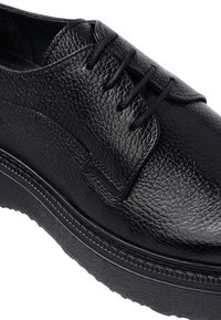 Zwarte leren dress shoe met textuur, ronde neus, zwarte veters en een stevige rubberen zool met geribbelde details.
