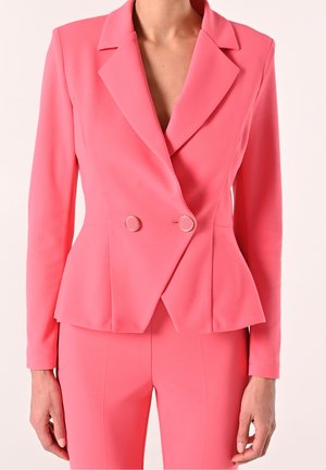 Frau trägt einen taillierten, doppelt geknöpften Blazer in Korallenrosa mit passenden, maßgeschneiderten Hosen, der Blazer ist mit goldfarbenen, runden Knöpfen versehen.