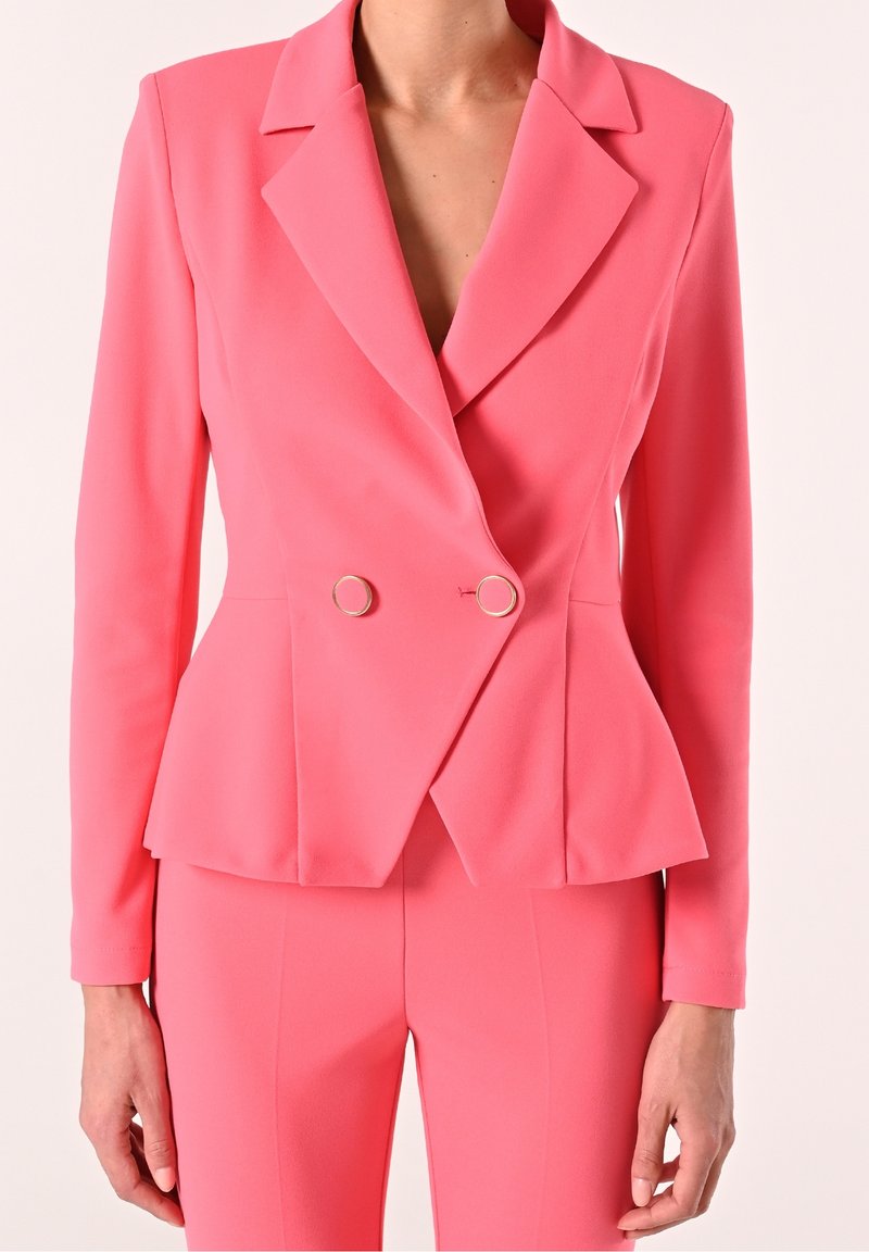 Femme portant un blazer double boutonnage rose corail ajusté avec un pantalon assorti et coupé sur mesure, avec des boutons circulaires dorés sur la veste.