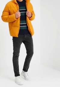 Veste matelassée jaune, pull bleu marine avec des rayures vertes et blanches, jean noir et chaussures blanches. Debout devant un fond clair.