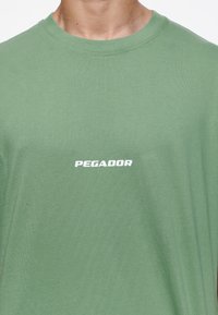 Detailní záběr na osobu v zeleném tričku s bílým textem "PEGADOR" umístěným ve středu na hrudi.
