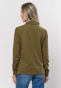 Oliivinvihreä pitkähihainen poolopaita, jossa on kaulus, sileä pinta ja hienovarainen sauma takana. Yhdistetty sinisiin farkkuihin.