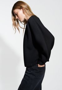 Maglione nero, oversize, con maniche ampie, realizzato in un tessuto testurizzato, abbinato a jeans scuri, mostrato di lato, evidenziando la silhouette.