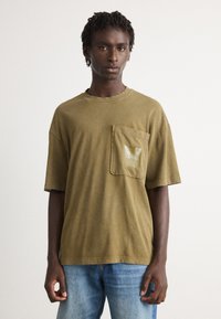 Ung mand med korte dreadlocks iført en overdreven olivengrøn t-shirt med lomme foran og blå jeans, stående mod en ensfarvet baggrund.