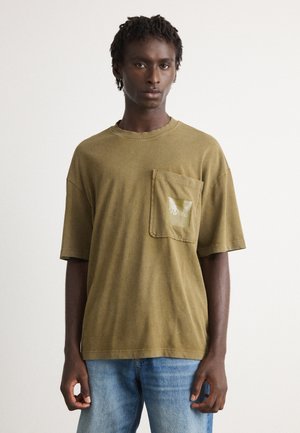 Young man with short dreadlocks wearing an olive green oversized t-shirt with a front pocket and blue jeans, standing against a plain background.