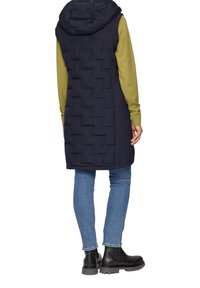 Marineblauer ärmelloser Daunenweste mit Kapuze, ausgestattet mit einem gesteppten Design und seitlichen Schlitzen. Kombiniert mit einem grünen Langarmshirt und blauen Jeans.