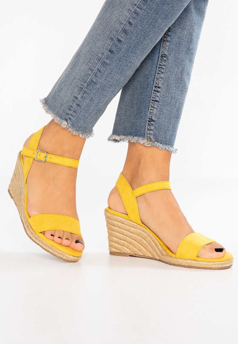 Pieds portant des sandales compensées jaunes avec des brides à la cheville et des orteils ouverts, associées à un jean bleu à ourlet effiloché.