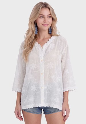 Donna bionda che indossa una camicetta bianca ricamata con fiori e bottoni, orecchini blu con nappe, abbinata a shorts di jeans, che guarda di lato.
