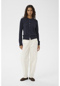 Marineblauer, gerippter Cardigan mit durchgehender Knopfleiste, langen Ärmeln und eng anliegenden Bündchen, kombiniert mit cremefarbenen, weit geschnittenen Hosen und braunen Slippern.