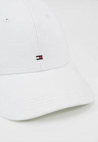 Hvid baseballkasket lavet af bomuld, med en buet skygge, seks ventilationshuller og et lille logo i rød, hvid og blå på siden.