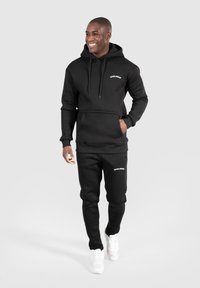 Hoodie noir avec cordons de serrage et poche avant, assorti à un pantalon de survêtement noir avec un logo, porté avec des baskets blanches.
