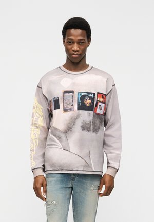 Férfi, aki hosszú ujjú, telefonos és fényképes mintával díszített pólót visel, világos, szakadt farmerrel, egyszerű háttér előtt áll.