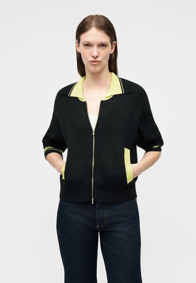 Γυναίκα που φοράει μαύρη ζακέτα με φερμουάρ, με κίτρινο γιακά και λεπτομέρειες στις τσέπες, τα χέρια στις τσέπες, συνδυασμένη με σκούρο μπλε τζιν, σε απλό φόντο.