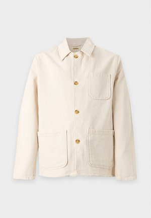 Jeune homme portant une veste beige boutonnée sur une chemise noire avec un pantalon noir et des chaussures habillées noires, debout avec une main dans la poche.