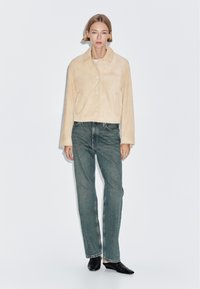 Donna che indossa una giacca beige con bottoni, jeans blu a gamba dritta e scarpe nere con punta, in piedi contro uno sfondo bianco uniforme.