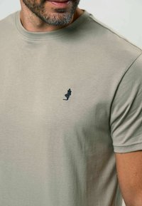 Gai smilškrāsas kokvilnas T-krekls ar apaļu dekoltē un īsām piedurknēm. Uz krūtīm ir mazs tumši zils izšūts logs.