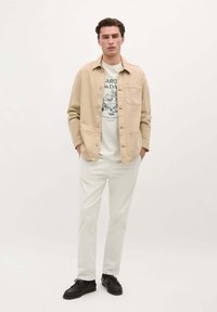 Beige knoopjesoverhemd-jack, crèmekleurig T-shirt met grafisch ontwerp, gecombineerd met witte broek en donkere schoenen. Glad stof, ontspannen pasvorm.