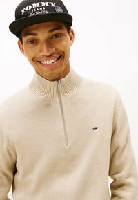 Μπεζ ζακέτα με φερμουάρ και ραμμένο γιακά, που διαθέτει μικρό κεντητό λογότυπο σε κόκκινο, άσπρο και μπλε. Φοριέται με μαύρο καπέλο μπέιζμπολ.