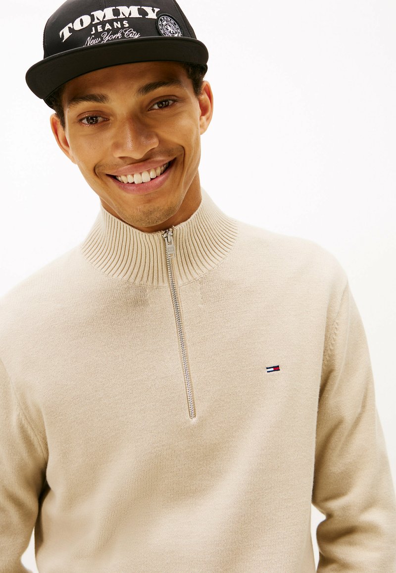 Suéter crema con cremallera y cuello acanalado, que presenta un pequeño logotipo bordado en rojo, blanco y azul. Llevado con una gorra de béisbol negra.