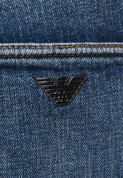 Το μπλε τζιν ύφασμα έχει αντίθετες πορτοκαλί ραφές. Ένα μαύρο μεταλλικό λογότυπο με στοιβαγμένα φτερά προβάλλει έντονα στην περιοχή της τσέπης.