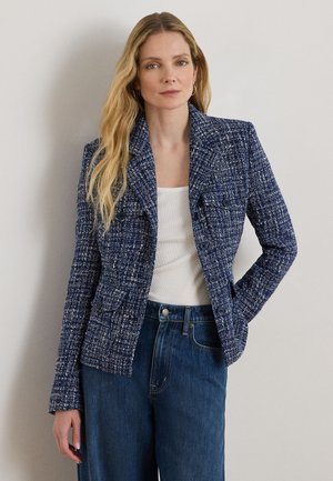 Kvinde med langt blondt hår iført en blå tekstureret blazer over en hvid top og blå højttaljede jeans, stående foran en neutral baggrund.