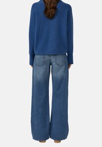 Vrouw gezien vanachter in een blauwe trui en wijdvallende vervaagde blauwe jeans met zakken en bruine schoenen.