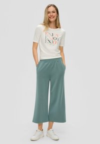 Witte graphic t-shirt met "ENJOY" in gekleurde letters; teal kleurige brede broek; witte sneakers. Zachte stof, verlopende pasvorm, zijzakken.