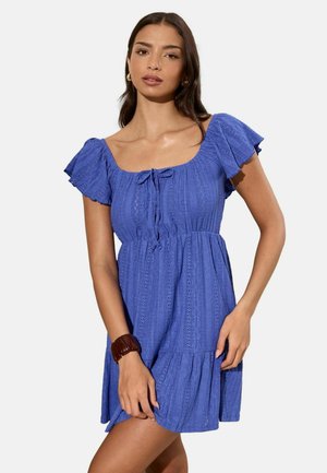 Junge Frau mit langen dunklen Haaren, die ein kurzärmeliges, strukturiertes blaues Kleid und ein dunkles Armband trägt, posiert vor einem schlichten hellen Hintergrund.