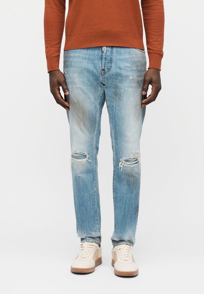 Homme portant un jean clair déchiré, des baskets blanches à semelles en gomme, et un pull à manches longues couleur orange brûlé, debout devant un fond blanc.