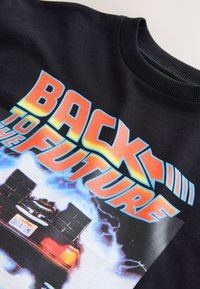 Hanorac negru cu un grafic mare multicolor al unei mașini DeLorean și textul "BACK TO THE FUTURE" deasupra, conturat în portocaliu și albastru aprins.
