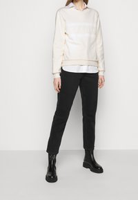 Ljus krämfärgad sweatshirt med präglad "CECILIE" logga, lager på lager över en vit skjorta med krage. Kombineras med svarta tapered jeans och svarta anklarstövlar.
