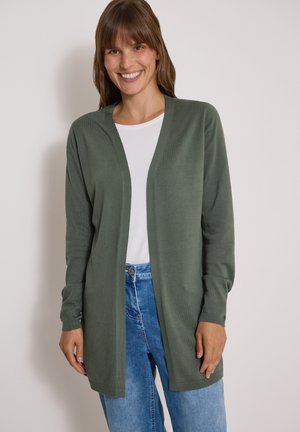 Smilende kvinde iført en lang olivengrøn cardigan med åbent front, hvid skjorte og blå jeans mod en ensfarvet baggrund.