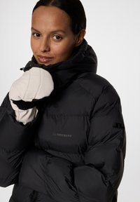 Svart pufferjacka med hög krage, med kviltad textur och en logotyp på bröstet. Händerna är täckta med gräddfärgade handskar.