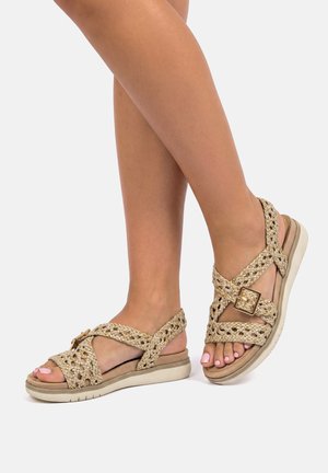 Pies con sandalias beige de tiras trenzadas con una hebilla dorada y suela blanca acolchada, mostrando las uñas de los pies pulidas sobre un fondo liso.