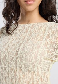 Woman with shoulder-length dark curly hair wearing a sheer lace cream top, showing details of the fabric and neckline.