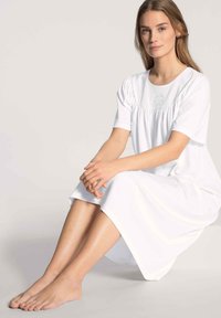 Camicia da notte bianca, lunga fino al ginocchio, in morbido tessuto con maniche corte e dettagli arricciati sul busto. Texture liscia e design semplice.