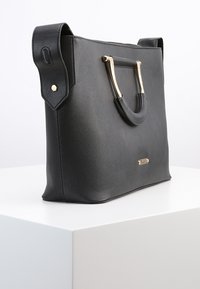 Svart syntetisk tote-väska med strukturerad yta, handtag i guldton och vinklad design. Har två topphandtag och sidodetaljer.