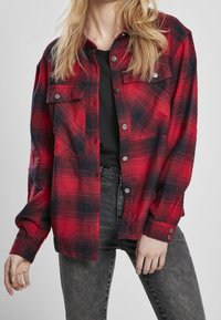 Kvinde iført en rød og sort ternet skjorte med knapper over en sort top og slidte sorte jeans, poserende med hænderne afslappet langs siden.